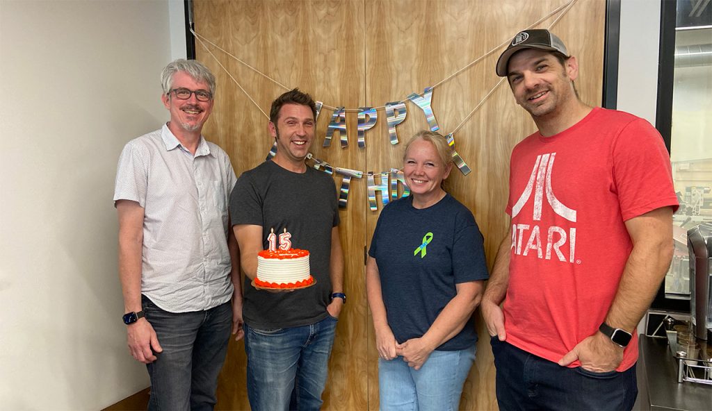 Pump Studios' four partners, (from Left to Right: Bard Collins, Peter Kaltenbach, Celeste Lamberth, and Kevin Keller). All four are standing in font of a wooden double door that has a holographic happy birthday sign. Peter us holding a cake that has a 15 lit candle.