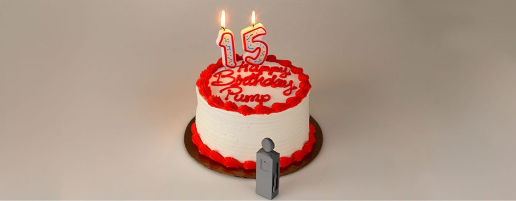 A red boarded birthday cake that reads "Happy Birthday Pump" There is a 1 and 5 candle lit up on the cake and a gray 3D printed gasoline pump in front of the cake.