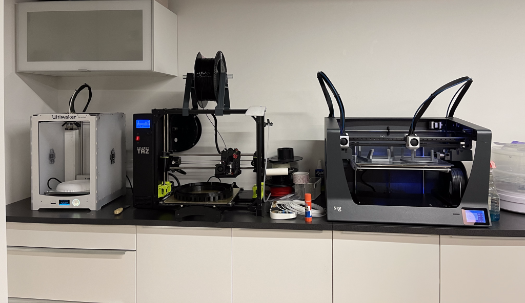 Three 3D printers and accessories. From the left to right: a white Ultimaker 2, a Lulzbot Taz and a sig 3D printer.