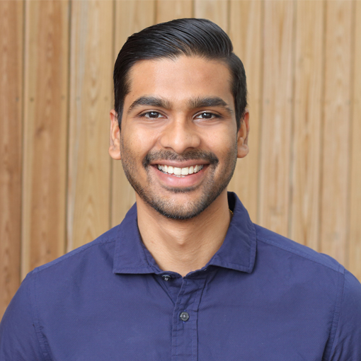 Parth Patel in a blue button up shirt, slicked back hair, and a big smile, standing in front of wood paneling.