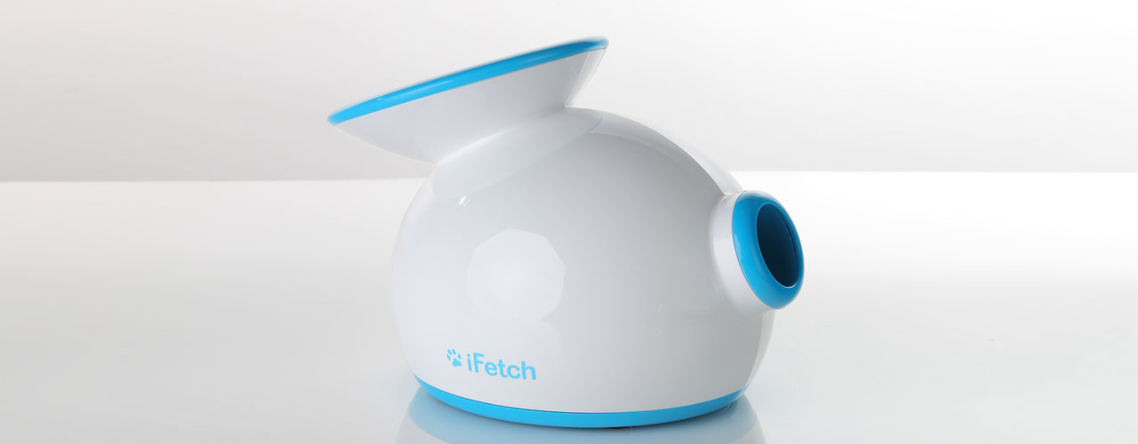 The first generation iFetch, a white and blue dog ball launcher with blue accents and blue logo, on a reflective table top.