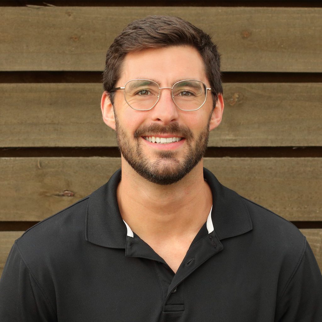 Greg Kinney in a black polo and glasses smiling in front of dark brown wood.