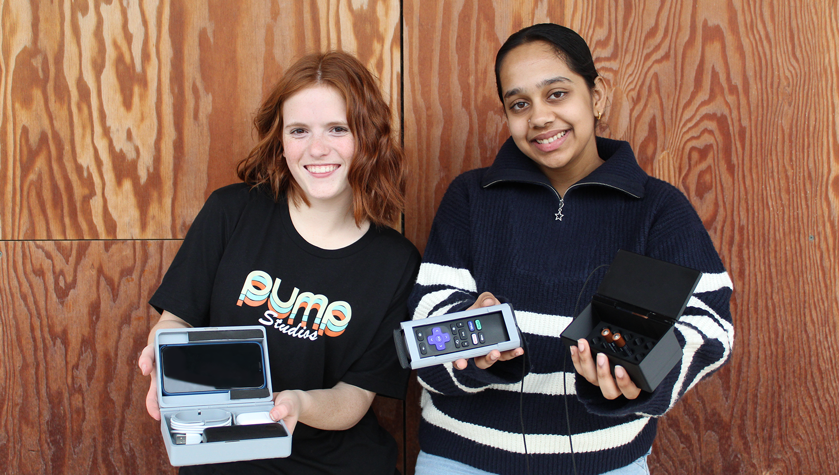 Junior interns Iris and Sivi standing with their 3D printed toolboxes in front of a wood paneled wall.