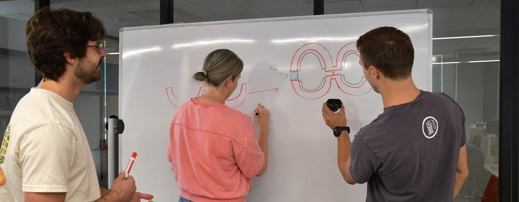 Liz, Jack and Greg standing in front of a whiteboard with a prototype and a drawing of the prototype.