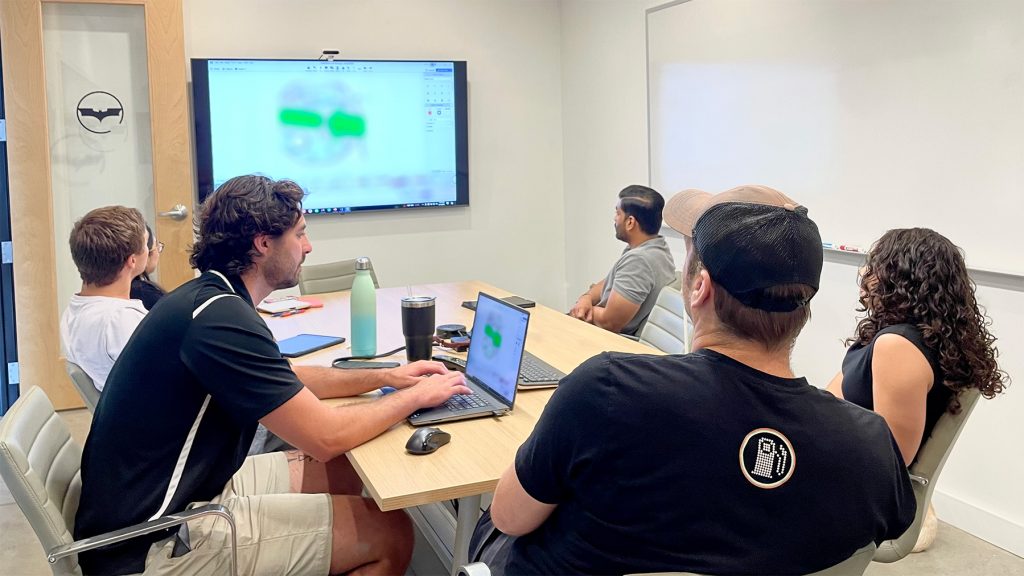 A group of engineers sit around a conference room table looking at a screen with a blurred image on the screen.