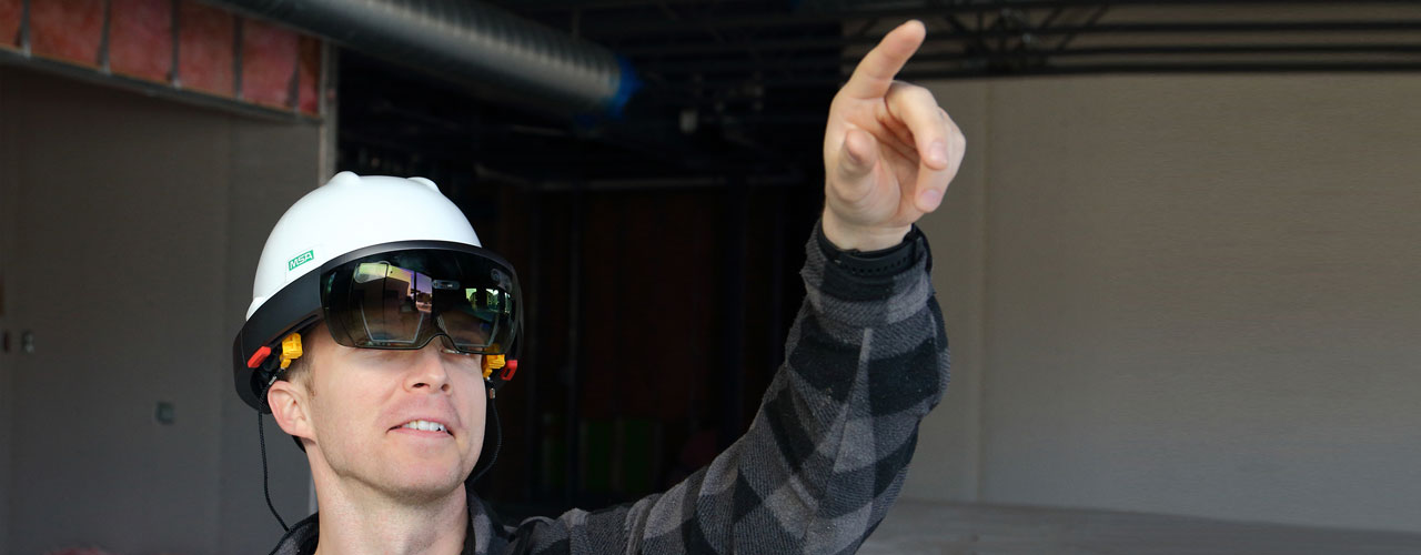 A man wearing a gray long sleeve flannel in an indoor construction site wearing the Trimble Connect for Hololens attached to a white hard hat.